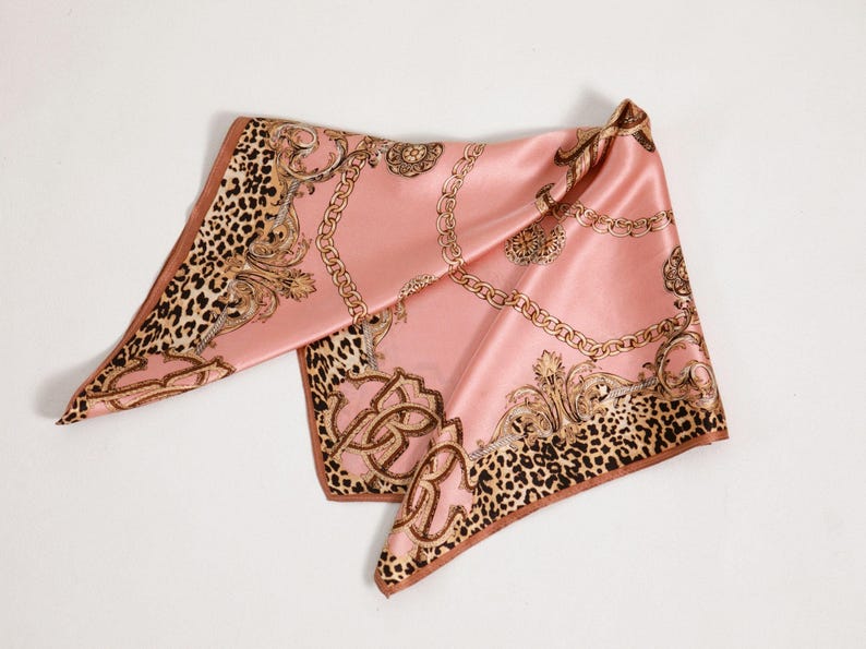 May include: A pink satin scarf with a gold chain and baroque design. The scarf has a leopard print border and a brown trim. The scarf is folded and displayed on a white surface.