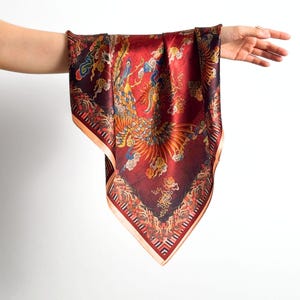 May include: A burgundy silk scarf with a detailed phoenix design in shades of gold, orange, and blue. The scarf has a patterned border and is held up by a hand against a white background. The scarf is square-shaped.