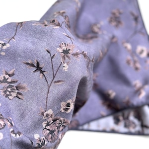 May include: A close-up of a lavender fabric with a floral pattern. The fabric has a subtle herringbone weave and features delicate brown and white flowers with green stems. The fabric is folded, creating depth and texture.