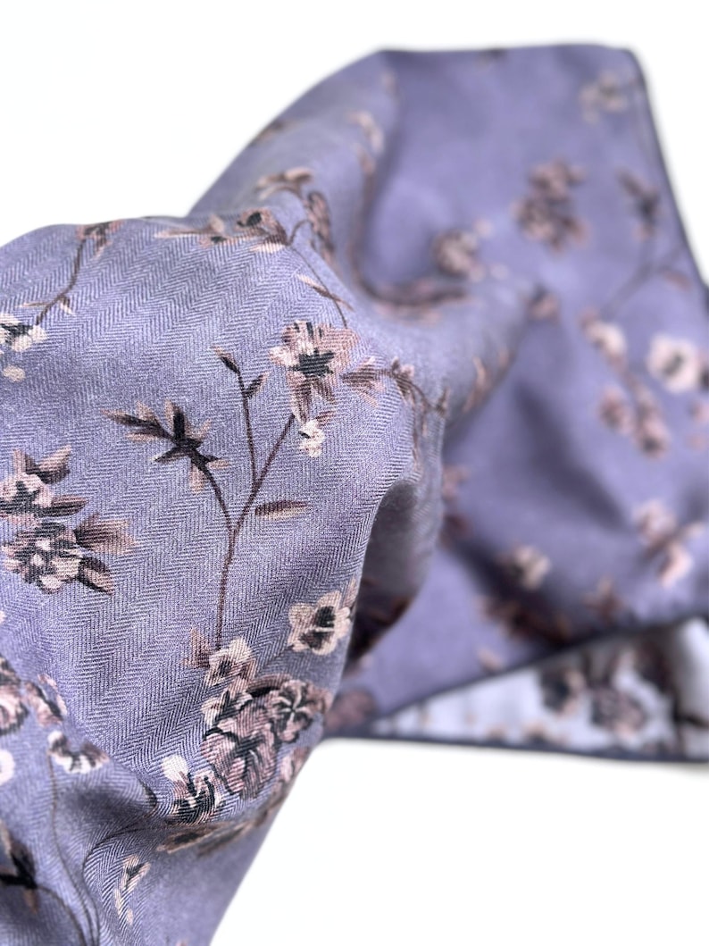 May include: A close-up of a lavender fabric with a floral pattern. The fabric has a subtle herringbone weave and features delicate brown and white flowers with green stems. The fabric is folded, creating depth and texture.