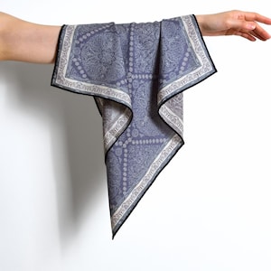 Könnte beinhalten: Ein blaues Paisley-Muster-Tuch mit schwarzem Rand wird von einer Hand gehalten. Das Tuch hat ein detailliertes Design mit weißen und grauen Akzenten. Das Tuch besteht aus einem weichen Material, wahrscheinlich Seide oder einem ähnlichen Stoff.