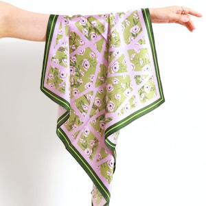 Peut inclure: Un foulard carré en soie avec un motif floral. Le foulard présente un motif floral vert clair et rose avec une bordure vert foncé. Le foulard est tenu par une main sur fond blanc.