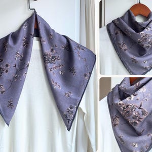 May include: A lavender scarf with a floral pattern. The scarf is displayed on a hanger, showcasing its square shape and dark trim. The floral design features various flower and leaf shapes.