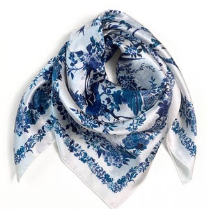 May include: A square scarf featuring a white base with a repeating blue floral and bird pattern. The design includes detailed illustrations of blooming flowers, leafy branches, and birds, framed by a decorative border of small floral motifs.