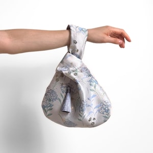 May include: A light gray fabric knot bag featuring a floral pattern with blue and green hydrangea blossoms and foliage. The bag has a single handle loop and a small, matching light gray tassel hanging from the knot at the top of the bag.