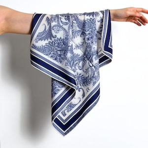May include: A white and blue paisley patterned scarf with a navy blue border. The scarf is held up by a person's arm, showcasing the intricate design and color contrast. The scarf's design features a detailed floral pattern.