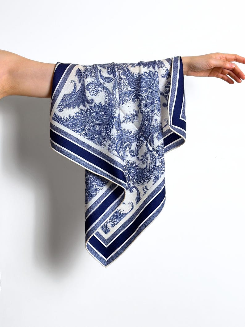 May include: A white and blue paisley patterned scarf. The scarf has a navy blue border with a striped design. The scarf is held up by a person's arm and hand, showcasing the fabric's drape and design. The scarf's pattern is a classic paisley.
