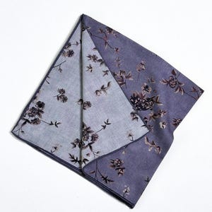 May include: A folded, square handkerchief with a floral pattern. The fabric is a muted purple with a lighter purple interior. The floral design features dark brown and black flowers and leaves.