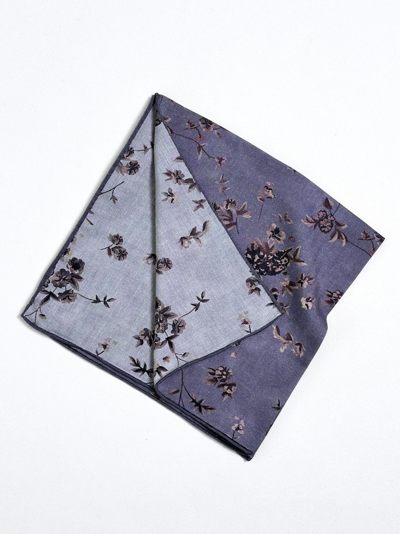 May include: A folded, square handkerchief with a floral pattern. The fabric is a muted purple with a lighter purple interior. The floral design features dark brown and black flowers and leaves.