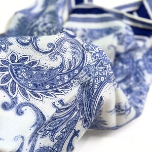 May include: A folded textile with a white background and a blue paisley pattern. The fabric has a detailed design with swirling floral and leaf motifs. The edges of the fabric are visible, showing a striped pattern in blue and white.