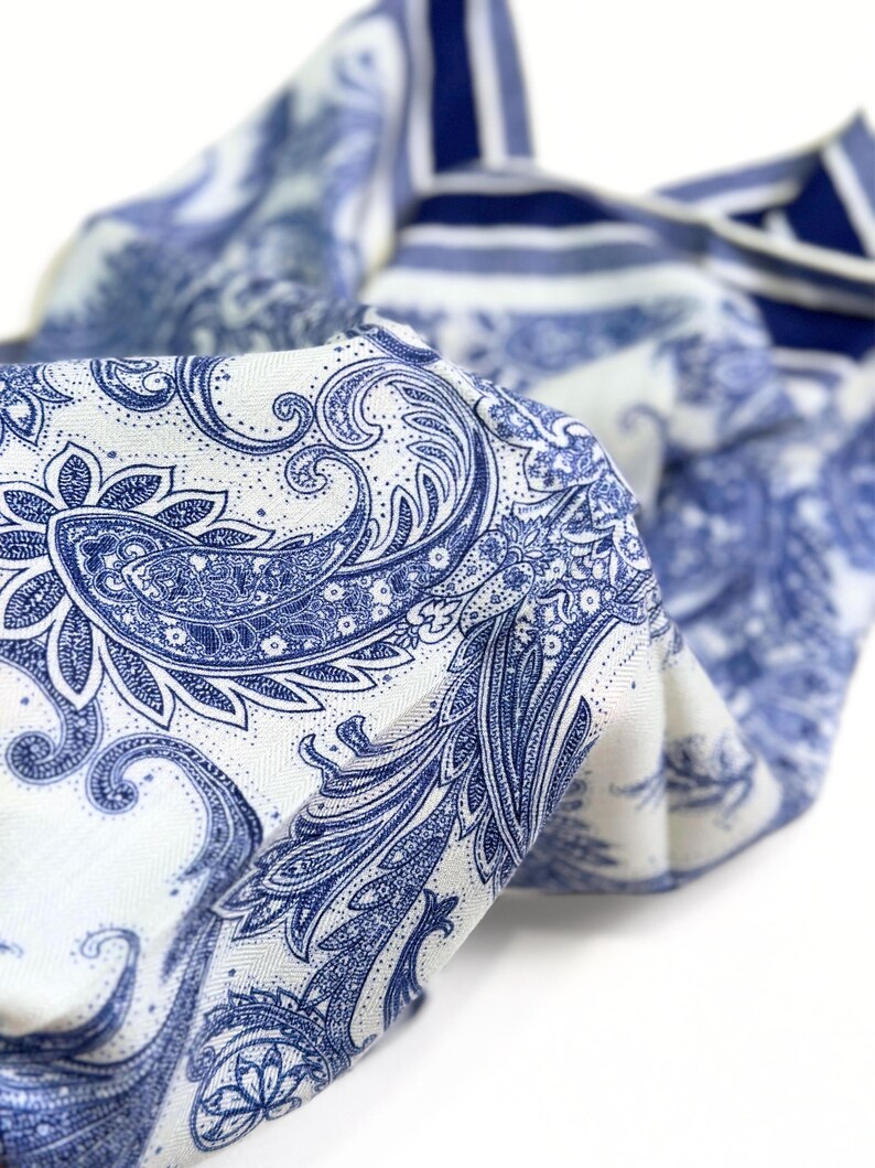 May include: A folded textile with a white background and a blue paisley pattern. The fabric has a detailed design with swirling floral and leaf motifs. The edges of the fabric are visible, showing a striped pattern in blue and white.