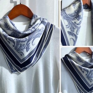 May include: A square scarf with a white background and a blue paisley design. The scarf has a dark blue border and is draped over a white sweater. The scarf is displayed on a wooden hanger.