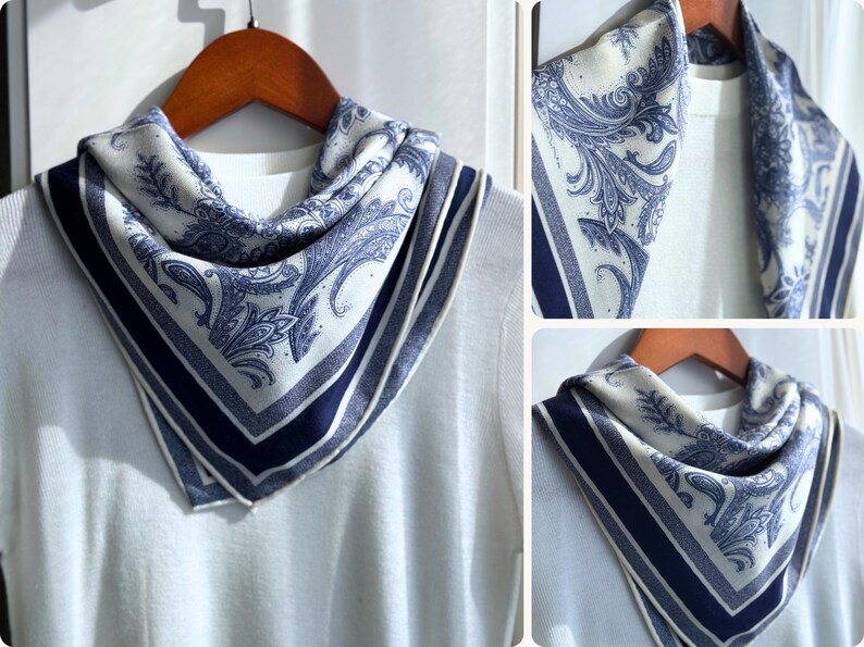 May include: A square scarf with a white background and a blue paisley design. The scarf has a dark blue border and is draped over a white sweater. The scarf is displayed on a wooden hanger.