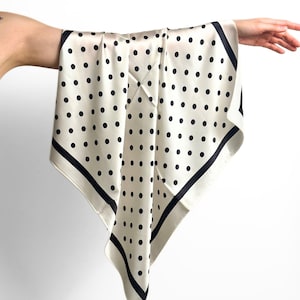 Puede incluir: Pañuelo cuadrado color crema con lunares negros y borde negro. El pañuelo se sostiene, mostrando su forma drapeada y textura sedosa. Un accesorio de moda versátil, adecuado para varios estilos.