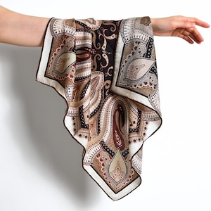 May include: A paisley patterned silk scarf in shades of brown, beige, and black. The scarf features a detailed design with a white border and a dark brown trim. The scarf is held up against a white background.
