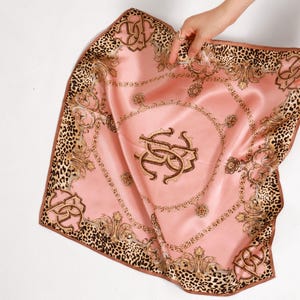 May include: A pink silk scarf with a leopard print border and ornate gold detailing. The center features a monogram design. The scarf is held up by two hands, showcasing its square shape and luxurious texture.