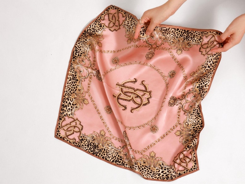 May include: A pink silk scarf with a leopard print border and ornate gold detailing. The center features a monogram design. The scarf is held up by two hands, showcasing its square shape and luxurious texture.