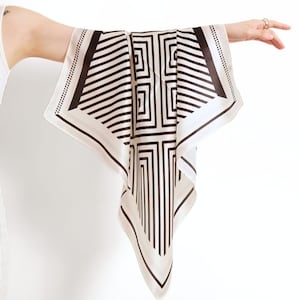 May include: A square, cream-colored scarf with a geometric black pattern. The design features a combination of straight lines and square shapes. The scarf is held up, showcasing its design and texture. The edges are finished with a dotted border.