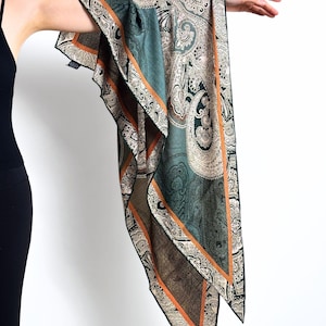 May include: A teal, brown, and cream paisley patterned scarf with an orange border. The scarf is draped over an arm against a white background. The scarf's design features intricate floral and teardrop motifs.