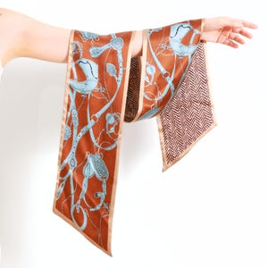 Op de afbeelding: Een lange, rechthoekige sjaal met een roestkleurige achtergrond en een lichtblauw paardensportthema. De sjaal heeft een beige rand en een bruin en beige visgraatpatroon aan één uiteinde.