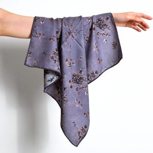 May include: A lavender-colored scarf with a floral pattern in shades of brown and black. The scarf has a dark border and is held up by a hand against a white background. The scarf is made of a soft, flowing fabric.