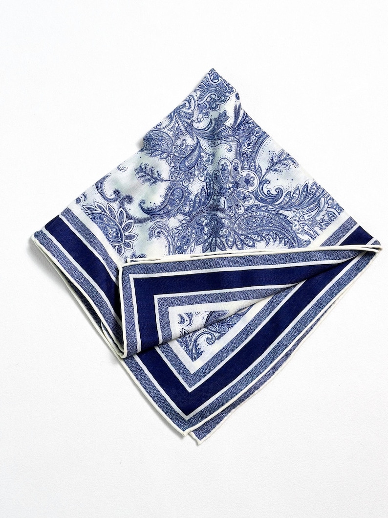 May include: A folded square handkerchief with a blue and white paisley pattern. The border features alternating stripes of white and navy blue, with a white trim. The handkerchief is made of a soft, possibly silk, material.