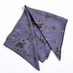 May include: A folded, lavender-colored pocket square with a floral pattern in shades of brown and black. The edges are trimmed with a darker navy blue. The fabric has a subtle sheen.