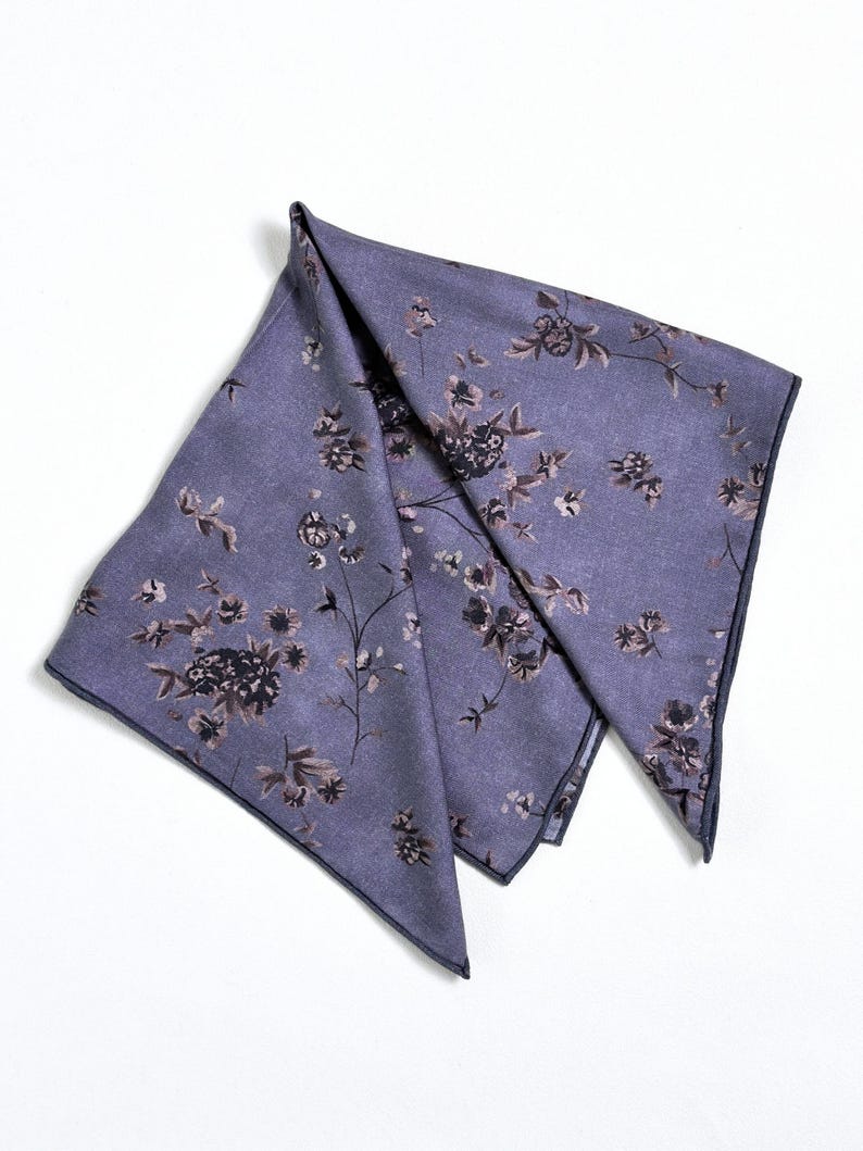 May include: A folded, lavender-colored pocket square with a floral pattern in shades of brown and black. The edges are trimmed with a darker navy blue. The fabric has a subtle sheen.