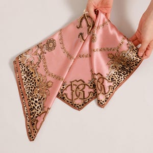May include: A pink silk scarf with a leopard print border and gold chain and scrollwork design. The scarf is being held up by two hands. The scarf has the letters "RR" in a gold monogram.