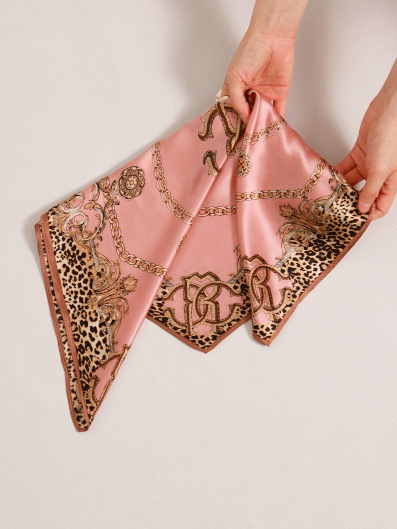 May include: A pink silk scarf with a leopard print border and gold chain and scrollwork design. The scarf is being held up by two hands. The scarf has the letters "RR" in a gold monogram.