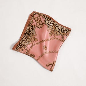 May include: A folded pink silk scarf with a brown border and a detailed pattern. The scarf features a leopard print section, gold chain and scrollwork designs, and a satin finish. The scarf is folded and lies on a white surface.
