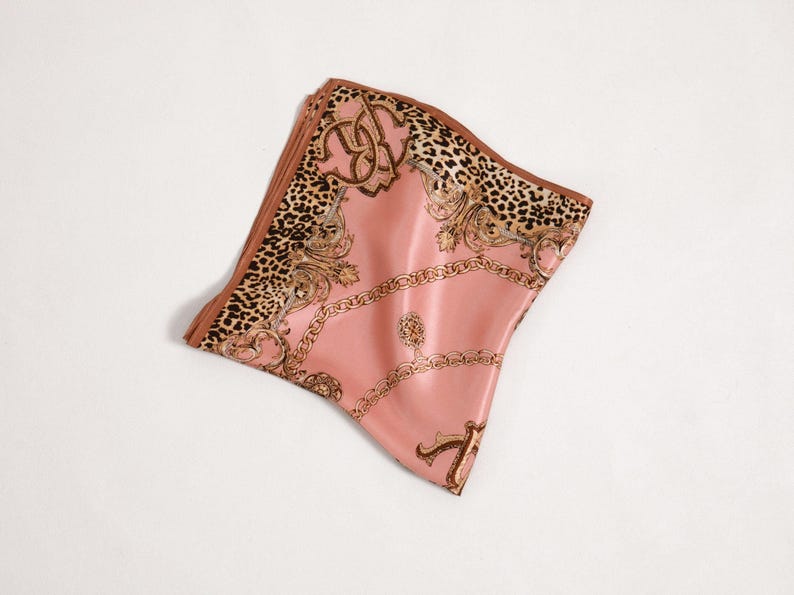 May include: A folded pink silk scarf with a brown border and a detailed pattern. The scarf features a leopard print section, gold chain and scrollwork designs, and a satin finish. The scarf is folded and lies on a white surface.