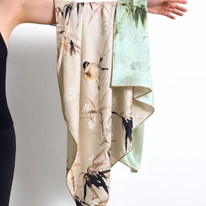 Può includere: Sciarpa in seta beige e verde chiaro con un motivo di uccelli e bambù. La sciarpa ha un bordo dorato ed è tenuta su uno sfondo bianco. La sciarpa è realizzata in un materiale morbido e fluido.
