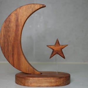May include: A wooden sculpture featuring a crescent moon and a star. The moon is a curved shape, and the star is a five-pointed design. Both are made of a warm-toned wood and are mounted on a round wooden base. The star is supported by a clear, thin rod.