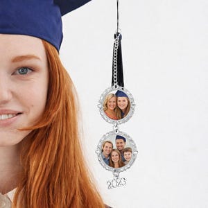 May include: A silver graduation ornament with two photo frames, each holding a picture. The frames are connected by a chain and a black tassel. The bottom frame has the year "2023" hanging below it. The ornament is hanging in front of a person wearing a graduation cap.
