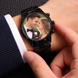 Peut inclure: Montre-bracelet noire avec une photo d'un couple sur le cadran. La montre a un bracelet en m&eacute;tal noir et un cadran rond. Le couple sur la photo sourit, peut-&ecirc;tre lors d'un mariage. La montre est port&eacute;e au poignet.