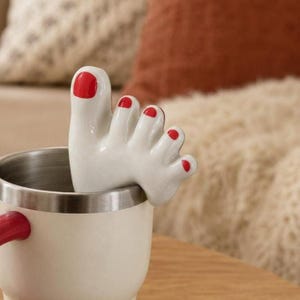 May include: A white ceramic foot-shaped mug accessory with red nail polish on the toes. The accessory is placed on the rim of a cream-coloured mug with a red handle. The mug has a silver interior.