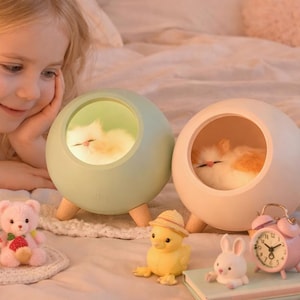 May include: Two cat-shaped night lights, one mint green and one off-white, each with a plush cat inside. The lights sit on wooden legs. A pink alarm clock, a yellow chick, a white bunny, and a pink teddy bear are also visible.