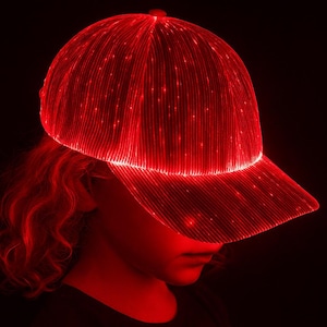 Peut inclure: Casquette de baseball rouge et lumineuse avec une visière incurvée. La casquette est illuminée de petites lumières, créant un motif rayé brillant. L'image est sur fond noir.