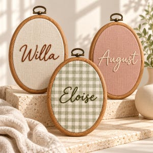 May include: Three oval embroidery hoops with wooden frames and bronze-colored hanging hardware. The left hoop features the name Willa in brown script on cream fabric. The center hoop shows Eloise in dark green script on green gingham fabric. The right hoop displays August in white script on dusty pink fabric.