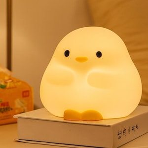 May include: A soft yellow night light shaped like a cartoon chick with black eyes and a yellow beak and feet. The light is sitting on a white book.