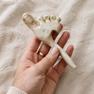 May include: A ceramic hand-shaped object with a thumb and four fingers, painted in off-white, pink, and green hues. The thumb and fingers have a textured appearance. The object is held in a hand with a gold ring.