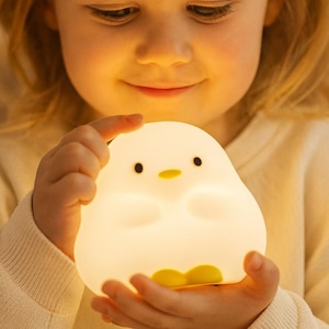 May include: A soft, glowing white night light shaped like a cartoon chick. The chick has black eyes and a yellow beak and feet. The night light is held by a child.