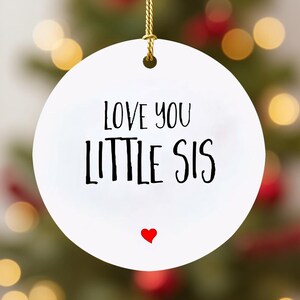 May include: A white, round ornament with the text "LOVE YOU LITTLE SIS" in black lettering. A small red heart is below the text. The ornament hangs from a gold string, perfect for holiday decor.