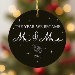 May include: Black round Christmas ornament with gold hanging string. The ornament has white text that reads "THE YEAR WE BECAME Mr & Mrs" with a diamond ring graphic and the year "2025". Small star graphics are scattered around the text.