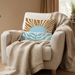 May include: A decorative throw pillow with a sun and sea design in shades of gold, blue, and white. The pillow is placed on a cream-colored armchair, draped with a beige knitted throw blanket.