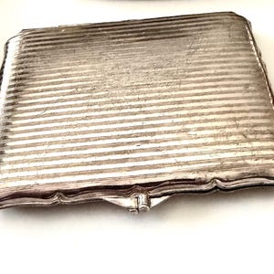 May include: A rectangular silver-toned metal cigarette case featuring a fine horizontal ribbed texture across the surface. The case has a scalloped edge design along the perimeter and a small central clasp mechanism on the front side for opening.