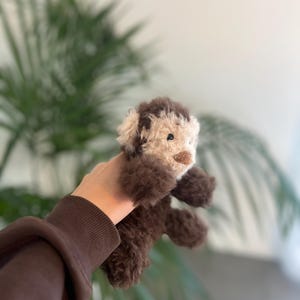 May include: A handmade plush toy dog with brown and cream-colored fur. The dog has a fluffy appearance, with a detailed face and small, dark eyes. The toy is held up against a blurred background of green foliage.