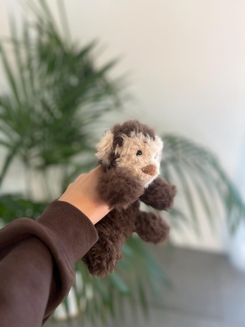 May include: A handmade plush toy dog with brown and cream-colored fur. The dog has a fluffy appearance, with a detailed face and small, dark eyes. The toy is held up against a blurred background of green foliage.