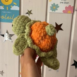 Peut inclure: Une tortue en peluche faite à la main, aux tons verts et oranges. La tête et les nageoires de la tortue sont vertes, tandis que sa carapace est orange vif. Le jouet est fait de fil doux et texturé, et est probablement un jouet pour enfants ou un objet décoratif.